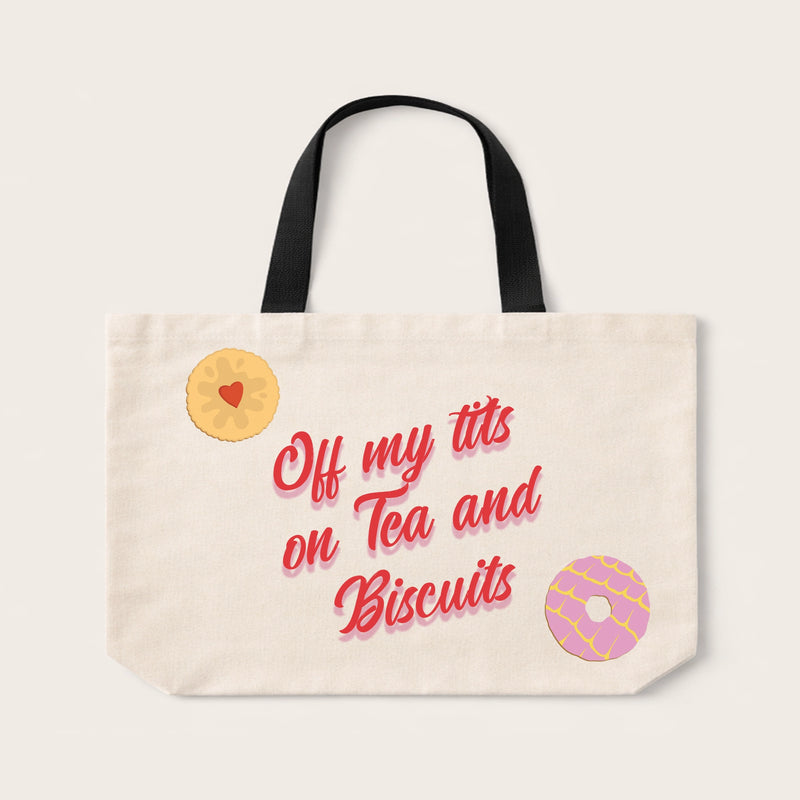 An off-white canvas tote bag with black shoulder straps, featuring a printed design with the phrase 'Off my tits on Tea and Biscuits' in red script, a yellow jam sandwich with a red heart, and a pink donut.
