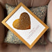 A personalized metallic foil heart-shaped art print with song lyrics, placed in a wooden frame, presented on a cushion.
