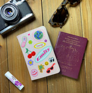 A pink personalised passport holder featuring a playful collage of stickers — smiley faces, cherries, palm trees, and a “Vacay” label — with the name Emily in bold lettering. The setup includes travel items such as sunglasses, a camera, lip balm, and a passport on a wooden background.
