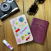 A pink personalised passport holder featuring a playful collage of stickers — smiley faces, cherries, palm trees, and a “Vacay” label — with the name Emily in bold lettering. The setup includes travel items such as sunglasses, a camera, lip balm, and a passport on a wooden background.