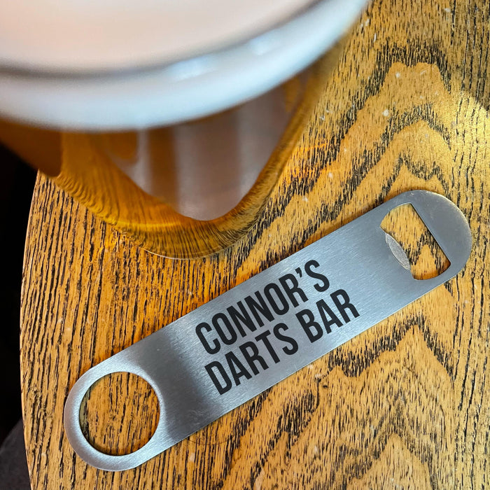 “Stainless steel bottle opener engraved with ‘Connor’s Darts Bar’ resting on a wooden bar top beside a pint glass.”