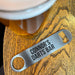 “Stainless steel bottle opener engraved with ‘Connor’s Darts Bar’ resting on a wooden bar top beside a pint glass.”