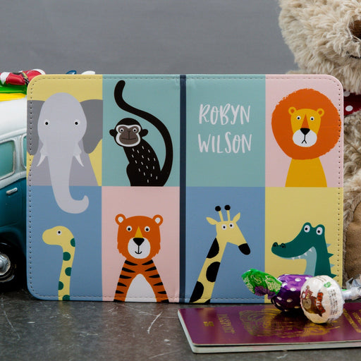 A children’s personalised passport holder with the name “Robyn Wilson” printed on the front. The cover features a grid of friendly illustrated safari animals — a lion, monkey, elephant, giraffe, tiger, and crocodile — in pastel colours. Props like a toy van, passport, and sweets create a playful travel-themed scene.