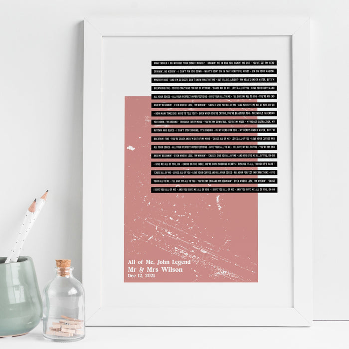 A framed lyric print with a dusty pink rectangular block on a beige background. The song lyrics “All of Me” by John Legend appear in black strip text above, with white lettering below reading: “All of Me, John Legend – Mr & Mrs Wilson – Dec 12, 2021.” The frame is white and sits on a light desk beside small jars and pencils.
