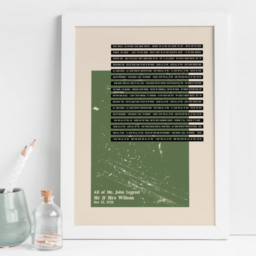 A framed lyric print with a minimalist beige background and a distressed green rectangular block. The lyrics of “All of Me” by John Legend are printed in narrow black strips across the top. At the bottom, white text reads: “All of Me, John Legend – Mr & Mrs Wilson – Dec 12, 2021.” The frame is white and sits on a light surface beside small glass jars and a pencil pot.