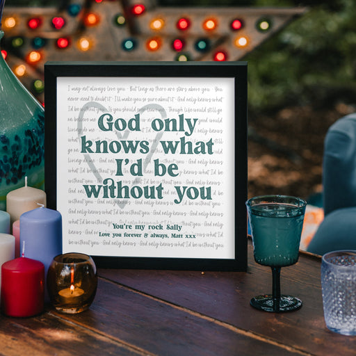 A framed print sits on a wooden outdoor table surrounded by colourful candles and glassware. The print reads in bold letters: “God only knows what I’d be without you,” with smaller text beneath saying “You’re my rock Sally. Love you forever & always, Matt xxx.” The background features blurred string lights and a cozy evening ambiance.