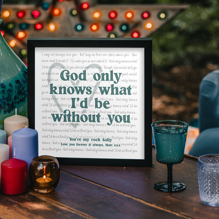 A framed print sits on a wooden outdoor table surrounded by colourful candles and glassware. The print reads in bold letters: “God only knows what I’d be without you,” with smaller text beneath saying “You’re my rock Sally. Love you forever & always, Matt xxx.” The background features blurred string lights and a cozy evening ambiance.