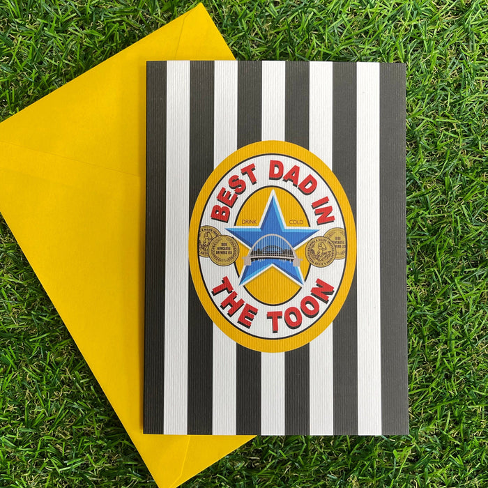 Black-and-white striped greeting card with “Best Dad in The Toon” badge design, shown with a yellow envelope on green grass background.