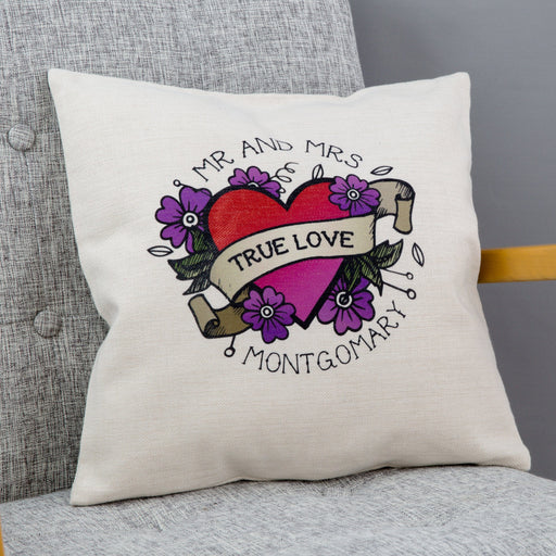 Cream cushion with tattoo-style heart and banner design reading “True Love” and personalised with “Mr and Mrs Montgomery”, placed on a grey chair.