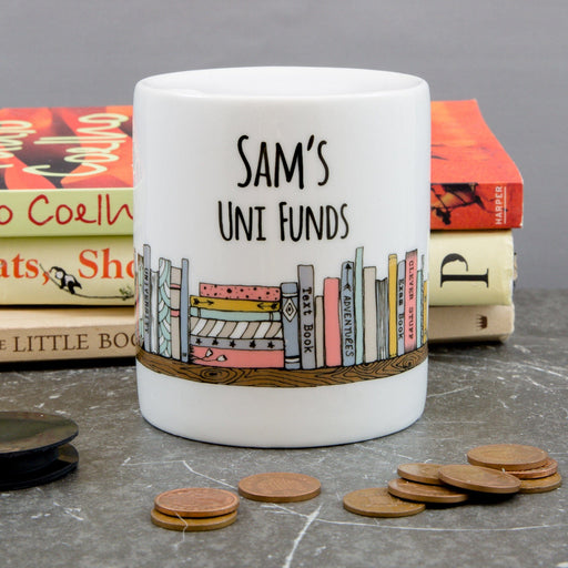 Personalised ceramic savings pot reading “Sam’s Uni Funds,” decorated with pastel book illustrations and shown on a grey tabletop with scattered coins.