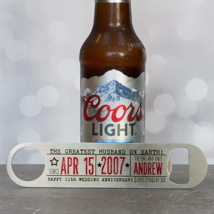 Personalised metal bottle opener engraved with “The Greatest Husband on Earth” and anniversary details, displayed in front of a Coors Light beer bottle on a grey surface.