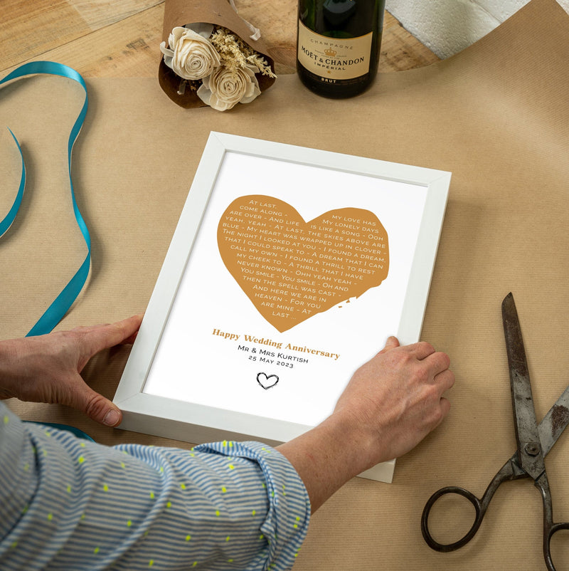 A person arranges a white-framed heart lyric print on a brown wrapping paper surface beside scissors, ribbon, flowers, and a bottle of Moët & Chandon champagne. The print has a gold heart containing the lyrics of “At Last,” with the inscription “Happy Wedding Anniversary – Mr & Mrs Kurtish – 25 May 2023” and a small hand-drawn heart symbol beneath.
