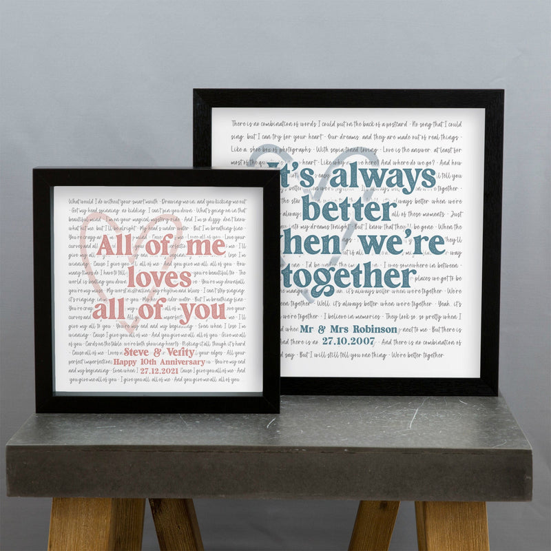 Two framed prints are displayed on a gray tabletop against a plain wall. The smaller frame on the left reads “All of me loves all of you” in pink text, with a personalized message below: “Steve & Verity — Happy 10th Anniversary 27.12.2021.” The larger frame on the right reads “It’s always better when we’re together” in blue text, with “Mr & Mrs Robinson — 27.10.2007” written below. Both prints have faint song lyrics or text in the background forming a heart shape.