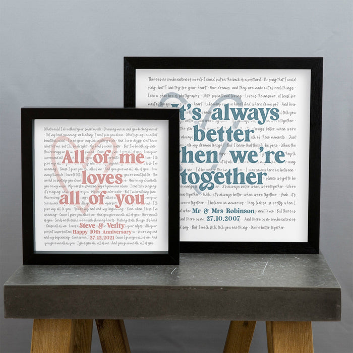 Two framed prints are displayed on a gray tabletop against a plain wall. The smaller frame on the left reads “All of me loves all of you” in pink text, with a personalized message below: “Steve & Verity — Happy 10th Anniversary 27.12.2021.” The larger frame on the right reads “It’s always better when we’re together” in blue text, with “Mr & Mrs Robinson — 27.10.2007” written below. Both prints have faint song lyrics or text in the background forming a heart shape.