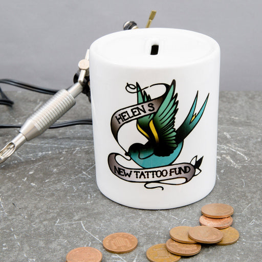 White ceramic money box with a tattoo-style swallow design and the text “Helen’s New Tattoo Fund,” shown with coins and a tattoo machine on a grey surface.