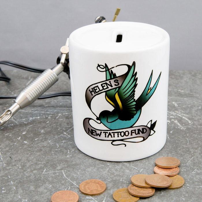 White ceramic money box with a tattoo-style swallow design and the text “Helen’s New Tattoo Fund,” shown with coins and a tattoo machine on a grey surface.