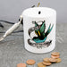 White ceramic money box with a tattoo-style swallow design and the text “Helen’s New Tattoo Fund,” shown with coins and a tattoo machine on a grey surface.