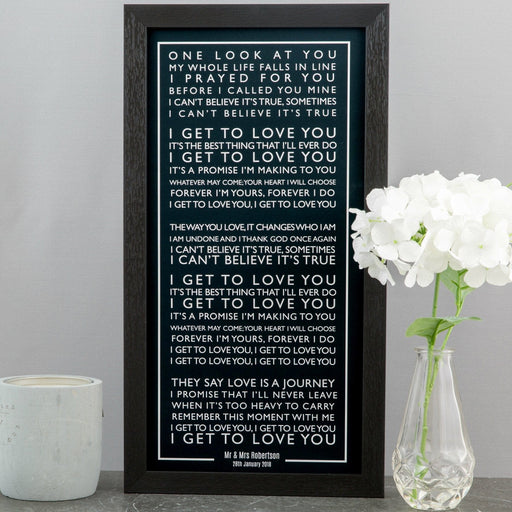 A black-framed lyric print with white text displayed on a gray wall beside a white candle and a vase of white flowers. The artwork features the lyrics “I Get to Love You” by Ruelle, with the words arranged in centered, uppercase text. At the bottom, it’s personalized with “Mr & Mrs Robertson – 28th January 2018.” The overall style is elegant and minimalist.