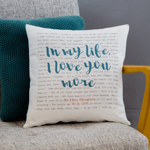 A square scatter cushion with personalized song lyrics printed on it, placed on a sofa with a yellow armchair in the background.