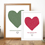 two framed heart-shaped lyric prints are displayed on a wooden surface. The larger print in a wooden frame features a green heart filled with song lyrics and the phrase “You’ll always be the better half of me” at the bottom. The smaller print in a white frame has a red heart with lyrics from “At Last,” and the message “Happy Valentine’s Day 2023 – Lucas & Monica” below. Both prints have small hand-drawn heart icons under the text.