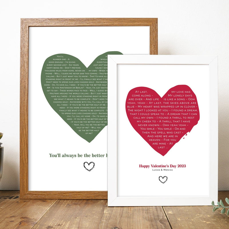 two framed heart-shaped lyric prints are displayed on a wooden surface. The larger print in a wooden frame features a green heart filled with song lyrics and the phrase “You’ll always be the better half of me” at the bottom. The smaller print in a white frame has a red heart with lyrics from “At Last,” and the message “Happy Valentine’s Day 2023 – Lucas & Monica” below. Both prints have small hand-drawn heart icons under the text.